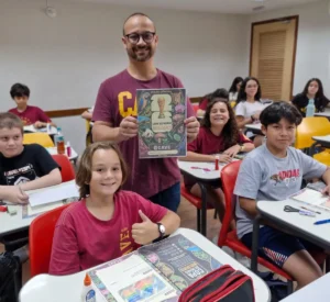 Professor Caique Duque e alunos do 7º ano do Colégio CAVE segurando o álbum de figurinhas científico Copa do Mundo Biológico" em sala de aula.